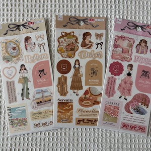 May include: Three sticker sheets with a variety of illustrations. The stickers feature drawings of women, bows, and decorative elements. The color palette includes beige, pink, and brown tones. Text includes "Radiant", "Dulcet", "Serenity", "Flowers bloom", and "Clearly".
