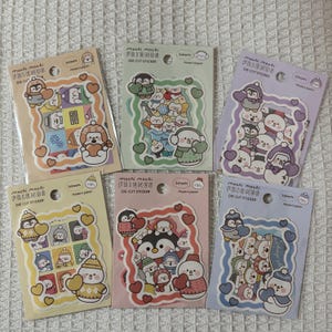 Puede incluir: Seis paquetes de pegatinas troqueladas con personajes de temática invernal, incluyendo pingüinos y muñecos de nieve. Cada paquete tiene un borde y un fondo de color diferente, con el texto "mochi mochi FRIENDS DIE CUT STICKER" y "Made in Japan".