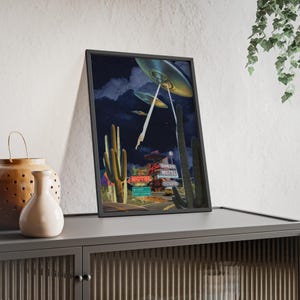 Könnte beinhalten: Ein gerahmter vertikaler Kunstdruck mit einer nächtlichen Wüstenlandschaft, hohen Saguaro-Kakteen, einem leuchtenden Sands Motel-Schild und mehreren fliegenden Untertassen am dunklen Himmel, die Lichtstrahlen auf den Boden unter sich werfen.
