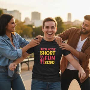 May include: A black t-shirt with the text "I'M NOT SHORT I'M FUN SIZED" in pink, yellow, and red. The shirt is being worn by a person, with two other people smiling and touching the person's shoulders. The image is taken outdoors.