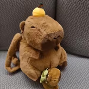 May include: A brown plush capybara toy with a soft, fuzzy texture. The capybara has closed eyes, a dark brown nose, and a small yellow fruit-shaped accessory on its head. A tiny green turtle charm is attached to the lower front of the plush body.