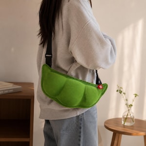 May include: A bright green, plush, pea pod-shaped crossbody bag with a soft, textured surface. The bag features a black adjustable strap with a plastic buckle and a small red fabric tag attached to the silver zipper pull on the side.