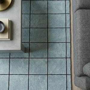 Peut inclure: Un tapis rectangulaire présentant une couleur de base bleu clair avec un motif de grille gris anthracite prononcé. Le tapis possède une surface à poils doux et texturés, posé sur un sol clair à côté d'un canapé gris et d'une table basse.