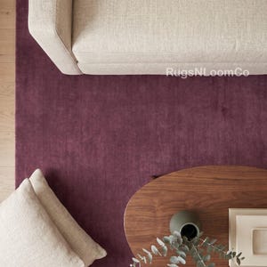 May include: A solid deep plum area rug with a smooth texture. The rug is positioned beneath a beige sofa, two beige cushions, and a dark wood oval coffee table holding a dark green vase with eucalyptus branches. Text reads RugsNLoomCo.