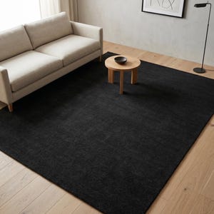 May include: A large, rectangular black rug dominates the foreground of a living room scene. A light-colored sofa and a small wooden side table with a dark bowl sit on the rug. The rug's texture is visible, adding depth to the room's decor.