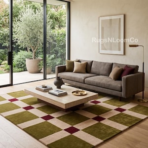Peut inclure: Un tapis rectangulaire présentant un motif géométrique en damier composé de carrés vert olive, crème et bordeaux foncé. Le tapis est placé sur un parquet clair sous une table basse rectangulaire et un canapé marron avec des coussins.