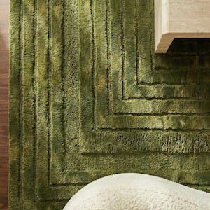 May include: A textured green rug with a geometric pattern. The rug has a rectangular shape with a series of vertical and horizontal lines. The rug is made of a soft, plush material. A white, rounded object is in the foreground.