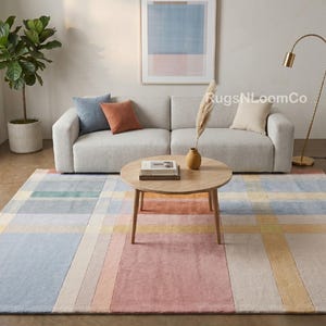 May include: A rectangular area rug featuring a grid pattern of intersecting lines in soft shades of blue, pink, yellow, and cream. The rug is positioned on a floor in front of a light gray sofa, a round wooden coffee table, and a floor lamp.