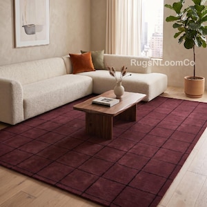 May include: A large rectangular area rug featuring a deep burgundy base colour with a prominent black grid pattern. The rug is positioned on a light-coloured wooden floor, covering a significant portion of the space beneath a wooden coffee table.