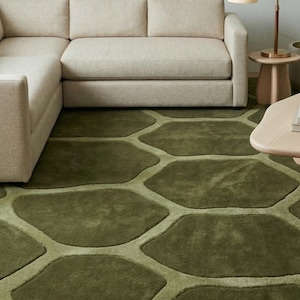 Puede incluir: Una alfombra verde con un diseño orgánico. La alfombra presenta una serie de formas irregulares interconectadas en diferentes tonos de verde. Un sofá seccional beige se encuentra sobre la alfombra, creando un espacio habitable acogedor.
