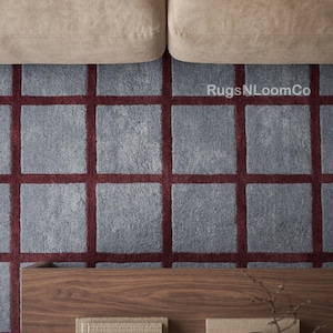 May include: A rectangular area rug featuring a grid pattern of light grey square sections separated by deep red lines. The text RugsNLoomCo is visible in the upper right quadrant of the rug. The rug is positioned beneath a beige sofa and a wooden table.