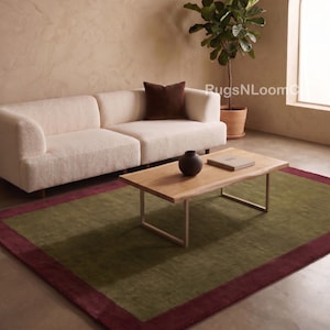 Peut inclure: Un tapis rectangulaire présentant un grand panneau central vert olive encadré par une large bordure bordeaux foncé. Le tapis est placé sur un sol lisse sous un canapé de couleur claire, une table basse en bois et une plante en pot.