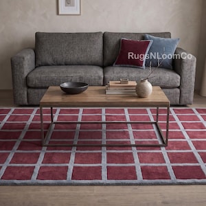 May include: A rectangular area rug featuring a grid pattern of deep red squares separated by light gray lines. The rug is positioned in front of a dark gray sofa, a wooden coffee table, and decorative items including a bowl and a vase.