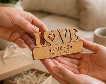 Puede incluir: Un letrero de madera cortado con láser que muestra la palabra LOVE con un recorte en forma de corazón en la letra O. Debajo, una base rectangular está grabada con la fecha 14 · 06 · 25 y los nombres EMILY & JACK en letra sans-serif.