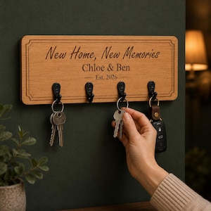 Puede incluir: Un soporte rectangular de madera para llaves con cuatro ganchos de metal negro. La madera está grabada con el texto New Home, New Memories, Chloe & Ben y Est. 2026. El tablero presenta un borde decorativo y está montado horizontalmente.