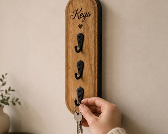 Puede incluir: Un soporte para llaves de madera vertical y arqueado con un acabado de veta de madera natural. Presenta la palabra Keys grabada en la parte superior, un pequeño símbolo de corazón debajo del texto y tres ganchos de metal negro en columna.