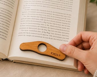 Puede incluir: Un sujetapáginas de madera de color marrón claro con una forma curva y suave y una abertura circular central. La madera presenta una veta natural y el nombre EMILY está grabado en una fuente oscura sin remates en el lado derecho del objeto.