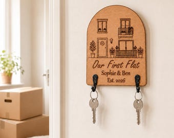 Puede incluir: Un llavero de pared de madera en forma de arco con una ilustración grabada con láser de una fachada de edificio. El texto dice Our First Flat, Sophie & Ben y Est. 2026. Dos ganchos de metal negro sostienen llaves plateadas en la madera.