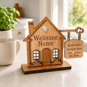 Puede incluir: Un letrero de madera con forma de casa sobre una base rectangular con el texto Welcome Home, un recorte de corazón y un letrero colgante sujeto por cadenas que dice Mia & Oliver, Our First Home y Est. 2026, con ventanas y follaje grabados.