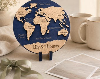 Puede incluir: Una placa circular de madera con un mapa del mundo sobre un fondo azul oscuro, continentes de madera clara y océanos etiquetados. El texto Lily & Thomas está grabado en la parte inferior. La placa se sostiene sobre un soporte de madera azul.