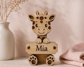 Puede incluir: Una figura de jirafa de madera de color marrón claro con un diseño recortado y manchas de color marrón oscuro. La jirafa sostiene un letrero rectangular grabado con el nombre Mia, flanqueado por pequeñas hojas, y se apoya en dos pies redondeados.