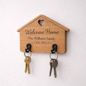 May include: Wooden house-shaped key holder with a heart cutout. It features the text "Welcome Home" and "The Williams Family - Est. 2023". Two sets of keys hang from black hooks. The wood has a natural grain.