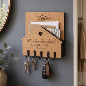 Puede incluir: Un soporte de pared de madera para correo y llaves con un panel frontal en forma de casa y un recorte de coraz&oacute;n. La madera est&aacute; grabada con el texto "Letters", "Where Our Story Begins", "The Miller Family" y "Est. 2026". Tiene cinco ganchos de metal negro.