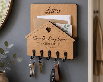 Puede incluir: Un soporte de pared de madera para correo y llaves con un panel frontal en forma de casa y un recorte de corazón. La madera está grabada con el texto "Letters", "Where Our Story Begins", "The Miller Family" y "Est. 2026". Tiene cinco ganchos de metal negro.