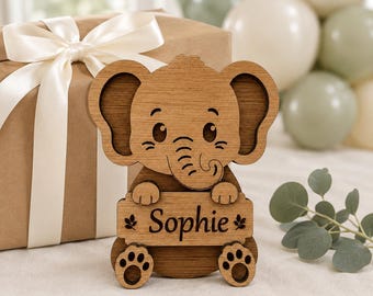 Puede incluir: Un recorte de madera marrón de un elefante de dibujos animados con orejas grandes y trompa, que sostiene un letrero rectangular grabado con el nombre Sophie y dos iconos de hojas pequeñas. Detalles de ojos y patas en marrón oscuro.