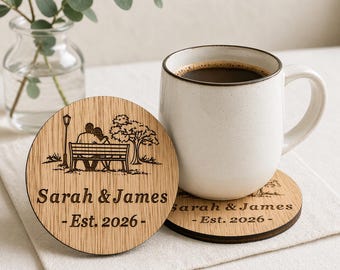 Puede incluir: Dos posavasos redondos de madera con un diseño grabado con láser de una pareja en un banco de parque, un árbol y una farola. Los posavasos tienen los nombres "Sarah & James" y el año "Est. 2026". Una taza blanca llena de café está sobre uno de ellos.