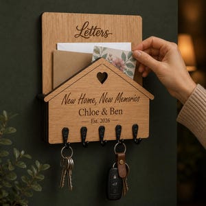 Puede incluir: Un organizador de correo y llavero de pared de madera con un panel frontal en forma de casa y un recorte de coraz&oacute;n. La madera est&aacute; grabada con el texto Letters, New Home, New Memories, Chloe & Ben y Est. 2026. Incluye cinco ganchos de metal negro.