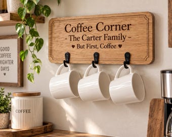 Puede incluir: Un soporte de pared rectangular de madera para tazas con tres ganchos de metal negro. La madera está grabada con el texto Coffee Corner, The Carter Family y But First, Coffee, junto a dos corazones. Tres tazas de cerámica blanca están colgadas.