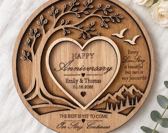 Puede incluir: Una placa circular de madera con un árbol cortado a láser, una forma de corazón central y siluetas de montañas. El texto dice: HAPPY Anniversary, Emily & Thomas, 01.06.2026, Every Love Story is beautiful but ours is my favourite, THE BEST IS YET TO COME, Our Story Continues.