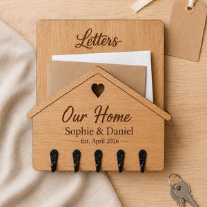 Puede incluir: Un organizador de correo y llavero de pared de madera clara con un panel frontal en forma de casa y un coraz&oacute;n recortado. La madera est&aacute; grabada con el texto "Letters", "Our Home", "Sophie & Daniel" y "Est. April 2026". Tiene cinco ganchos de metal negro.