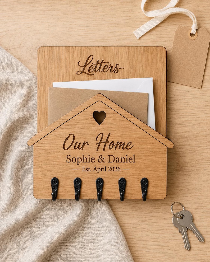 Puede incluir: Un organizador de correo y llavero de pared de madera clara con un panel frontal en forma de casa y un coraz&oacute;n recortado. La madera est&aacute; grabada con el texto "Letters", "Our Home", "Sophie & Daniel" y "Est. April 2026". Tiene cinco ganchos de metal negro.