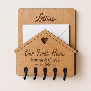 Puede incluir: Un organizador de correo y llavero de pared de madera marr&oacute;n claro con un panel frontal en forma de casa y un recorte de coraz&oacute;n. El texto dice Letters, Our First Home, Emma & Oliver y Est. 2026. Incluye cinco ganchos de metal negro.
