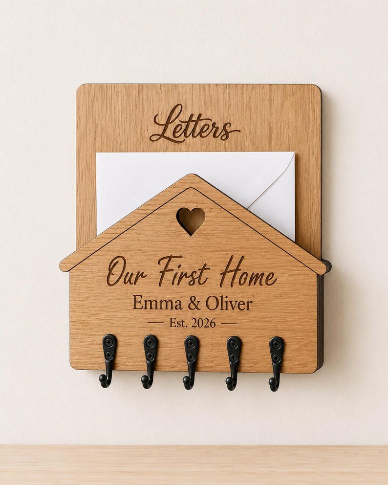 Puede incluir: Un organizador de correo y llavero de pared de madera marr&oacute;n claro con un panel frontal en forma de casa y un recorte de coraz&oacute;n. El texto dice Letters, Our First Home, Emma & Oliver y Est. 2026. Incluye cinco ganchos de metal negro.