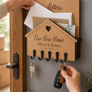 Puede incluir: Un organizador de correo y llavero de pared de madera clara con forma de casa. Cuenta con un recorte en forma de coraz&oacute;n, cinco ganchos de metal negro y texto grabado que dice Letters, Our New Home, Olivia & James y Est. 2026.