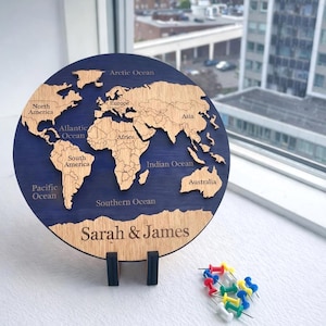 May include: A circular wooden world map with continents cut out, set against a dark blue backdrop. The map is labelled with ocean names and continents. The names "Sarah & James" are engraved at the bottom. Includes colourful push pins.