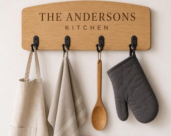 Puede incluir: Un estante de pared rectangular de madera clara con cuatro ganchos de metal negro. La madera está grabada con el texto THE ANDERSONS KITCHEN. Colgados de los ganchos hay un delantal beige, un paño de cocina a rayas beige, una cuchara de madera y un guante de horno gris oscuro.