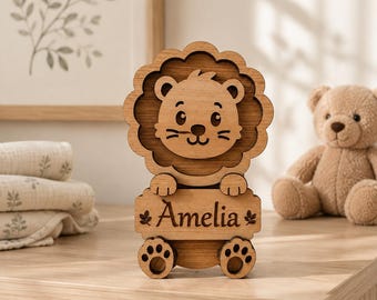 Puede incluir: Un recorte de madera de color marrón claro con forma de león y melena festoneada. El león sostiene una placa rectangular grabada con el nombre Amelia, flanqueada por dos pequeños iconos de hojas. El diseño incluye huellas de patas recortadas.