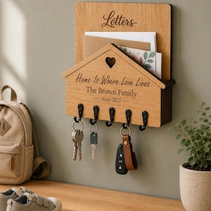 Puede incluir: Un organizador de correo y llavero de pared de madera clara con cinco ganchos de metal negro. El panel frontal tiene forma de casa con un recorte de coraz&oacute;n y el texto grabado Letters, Home Is Where Love Lives, The Brown Family, Since 2022.