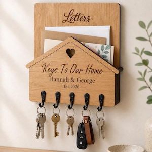 Puede incluir: Un organizador de correo y llavero de pared de madera clara. La secci&oacute;n superior tiene la etiqueta Letters. El frente en forma de casa presenta un recorte de coraz&oacute;n, el texto Keys To Our Home, Hannah & George y Est. 2026, con cinco ganchos negros.
