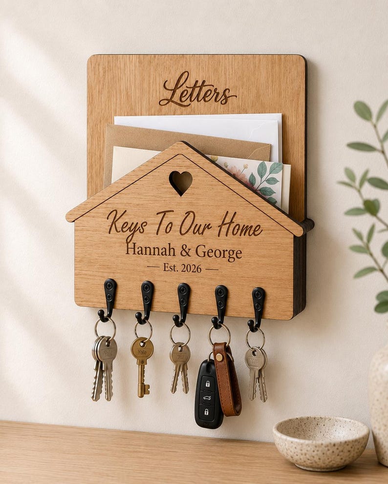 Puede incluir: Un organizador de correo y llavero de pared de madera clara. La secci&oacute;n superior tiene la etiqueta Letters. El frente en forma de casa presenta un recorte de coraz&oacute;n, el texto Keys To Our Home, Hannah & George y Est. 2026, con cinco ganchos negros.