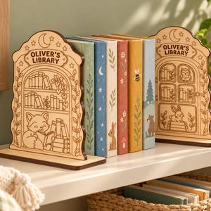 Puede incluir: Dos sujetalibros de madera clara con diseños grabados de estanterías, un zorro leyendo y un búho. Cada sujetalibros es arqueado con el texto "OLIVER'S LIBRARY" en la parte superior. Sostienen cinco libros coloridos con ilustraciones de naturaleza.