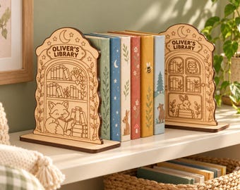Puede incluir: Dos sujetalibros de madera clara con diseños grabados de estanterías, un zorro leyendo y un búho. Cada sujetalibros es arqueado con el texto "OLIVER'S LIBRARY" en la parte superior. Sostienen cinco libros coloridos con ilustraciones de naturaleza.