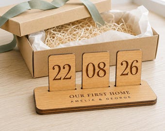 Puede incluir: Un calendario de escritorio de madera marrón claro con tres placas rectangulares verticales que muestran la fecha 22 08 26. La base está grabada con el texto OUR FIRST HOME AMELIA & GEORGE. Se presenta con una caja de regalo de cartón y cinta verde.