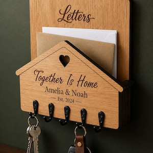 Puede incluir: Un organizador de correo y llavero de pared de madera clara. La secci&oacute;n superior est&aacute; grabada con la palabra Letters. La secci&oacute;n inferior en forma de casa presenta un recorte de coraz&oacute;n, el texto Together Is Home, Amelia & Noah, Est. 2024 y cinco ganchos de metal negro.