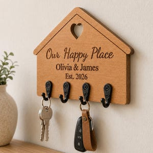 Puede incluir: Un soporte para llaves de madera con forma de casa y cuatro ganchos de metal negro. La madera está grabada con un recorte en forma de corazón en la parte superior y el texto "Our Happy Place", "Olivia & James" y "Est. 2026". Dos llaves cuelgan.