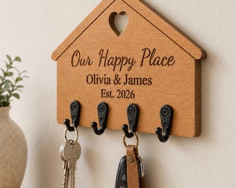 Puede incluir: Un soporte para llaves de madera con forma de casa y cuatro ganchos de metal negro. La madera está grabada con un recorte en forma de corazón en la parte superior y el texto "Our Happy Place", "Olivia & James" y "Est. 2026". Dos llaves cuelgan.